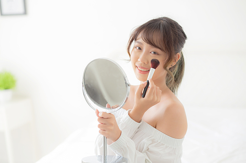 Beauty portrait young asian woman smile with face looking mirror applying makeup with brush cheek in the bedroom, beautiful of girl holding blusher, skin care and cosmetic concept.