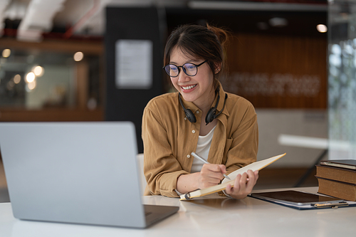 Portrait of young asian woman using laptop and writing making list taking notes in notepad working or learning on laptop indoors- educational course or training, seminar, education online concept