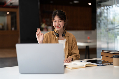 Joyful young asian female in wireless headphones waving wand greeting to screen while sitting at table and having video chat with business partners using laptop against of comfortable office.