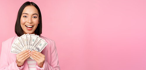 Finance, microcredit and people concept. Happy smiling asian businesswoman showing dollars money, standing in suit against pink background.