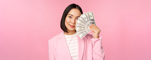 Microcredit, investment and business people concept. Young asian businesswoman, corporate lady showing money, cash dollars, smiling pleased, pink background.