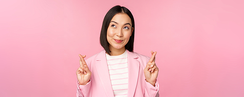 Happy lucky businesswoman, asian corporate lady wishing, making wish, hoping for smth and praying, standing in suit over pink background.