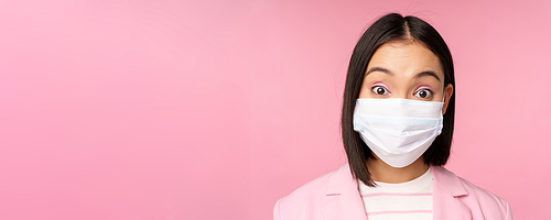 Close-up portrait of asian businesswoman in medical face mask, looking surprised, standing in suit over pink background. Copy space