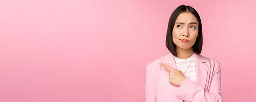 Doubtful business woman, sulking and grimacing while pointing, looking left at banner with skeptical face expression, posing against pink background.