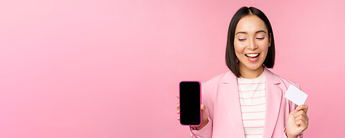 Smiling korean businesswoman in suit, showing mobile phone screen, credit card, showing online banking application interface, pink background.