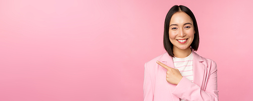 Portrait of asian business woman, saleswoman in suit pointing finger left, showing banner advertisement, smiling and looking professional, pink background.