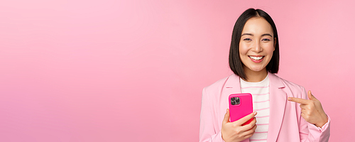 Portrait of smiling asian businesswoman pointing at her mobile phone, recommending smartphone app, application on cellphone, standing over pink background.