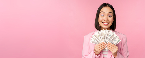 Finance, microcredit and people concept. Happy smiling asian businesswoman showing dollars money, standing in suit against pink background.