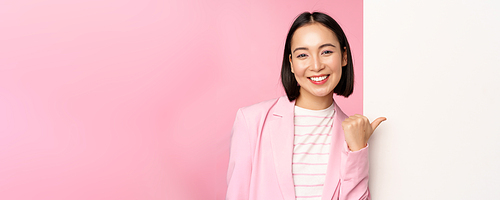 Portrait of smiling asian businesswoman in suit, corporate lady pointing finger at white empty wall, board with info or advertisement, standing over pink background.