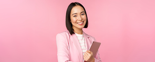 Professional smiling asian businesswoman, standing with digital tablet, wearing suit for office work, looking confident and happy, posing against pink background.