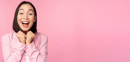 Portrait of happy asian office lady, ceo businesswoman in suit rejoicing and laughing, winning, celebrating, achieve goal and rejoice, standing over pink background.