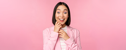 Happy young office lady, korean business woman wearing suit, looking surprised at camera, posing against studio background in pink.