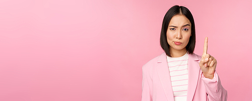 Asian businesswoman with serious, concerned face expression, showing stop motion, taboo, prohibit gesture, disapprove smth bad, standing over pink background.