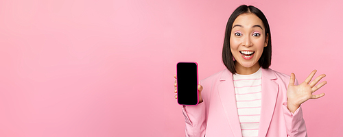 Surprised, enthusiastic asian businesswoman showing mobile phone screen, smartphone app interface, standing against pink background.