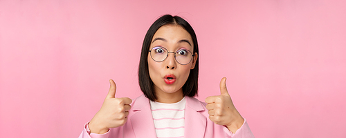 Close up portrait of impressed corporate woman, asian business lady in glasses, showing thumbs up, looking amazed at camera, recommending, pink background.