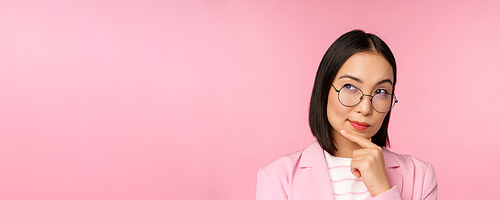 Korean businesswoman thinking, wearing glasses, looking thoughtful at camera, making decision, standing over pink background.