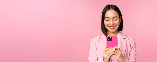 Portrait of smiling business woman, asian corporate person using smartphone, mobile phone application, standing over pink background.