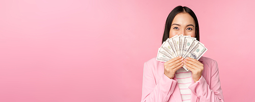 Happy asian lady in suit holding money, dollars with pleased face expression, standing over pink background. Copy space