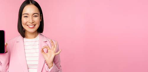 Satisfied smiling asian businesswoman recommending mobile phone app, website company on smartphone, showing screen and okay sign, pink background.