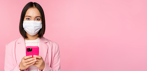 Business people and covid-19 concept. Japanese corporate office lady in suit and medical face mask, using mobile phone and smiling at camera, pink background.