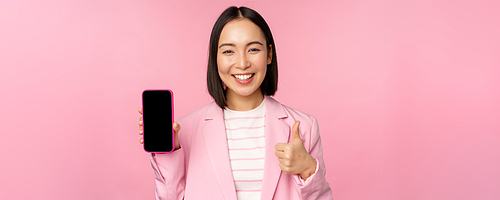 Satisfied smiling asian businesswoman recommending mobile phone app, website company on smartphone, showing screen and thumbs up, pink background.