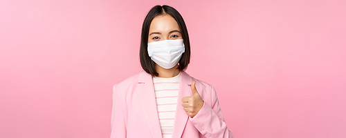 Asian businesswoman in suit and medical face mask, showing thumbs up, recommending wearing personal protective equipment in office during covid-19 pandemic, pink background.