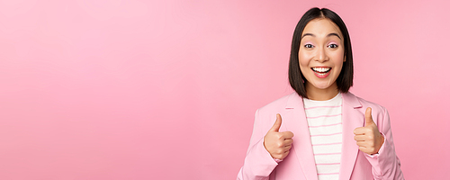 Portrait of asian businesswoman smiling satisfied, showing thumbs up, praise, like and approve, standing in suit over pink background.