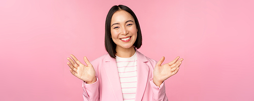 Portrait of smiling asian office lady, businesswoman raising hands, waving and looking happy, posing in suit against pink background.