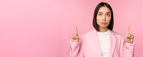 Upset asian corporate woman in office suit, pointing and looking up with disappointed face expression, standing over pink background.