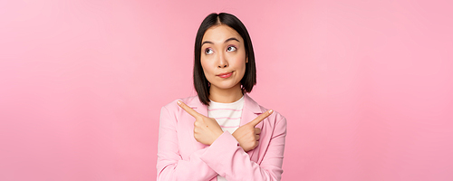 Decision concept. Young asian businesswoman, corporate worker pointing fingers sidways, two directions, showing variants, looking indecisive while choosing, pink background.