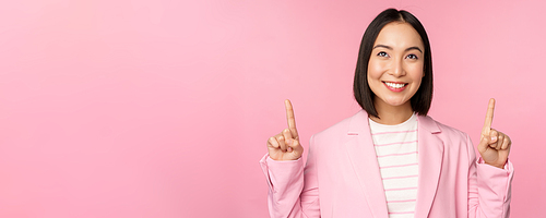 Smiling korean businesswoman, pointing fingers up, showing advertisement, banner or logo on top, standing in suit over pink background.