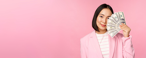 Microcredit, investment and business people concept. Young asian businesswoman, corporate lady showing money, cash dollars, smiling pleased, pink background.