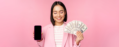 Image of korean successful corporate woman showing money, dollars and smartphone app screen, interface of mobile phone application, concept of investment and finance, pink background.