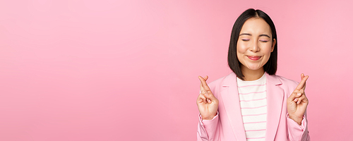 Happy lucky businesswoman, asian corporate lady wishing, making wish, hoping for smth and praying, standing in suit over pink background.