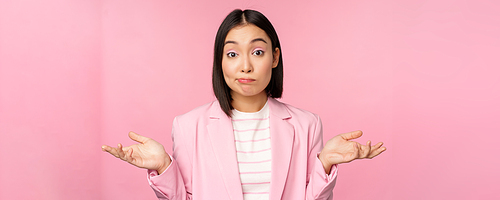 Portrait of confused asian businesswoman shrugging shoulders, looking clueless and puzzled, dont know, cant say, standing over pink background in office suit.