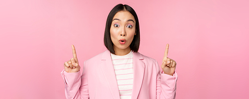 Portrait of asian businesswoman, corporate worker in suit, looks surprised by advertisement, points finger up, showing banner or logo on top, stands over pink background.