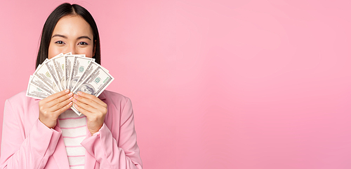 Happy asian lady in suit holding money, dollars with pleased face expression, standing over pink background. Copy space