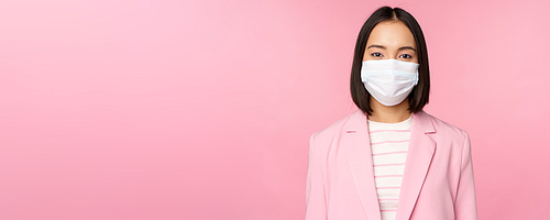 Portrait of asian businesswoman in medical face mask, wearing suit, concept of office work during covid-19 pandemic, standing over pink background.