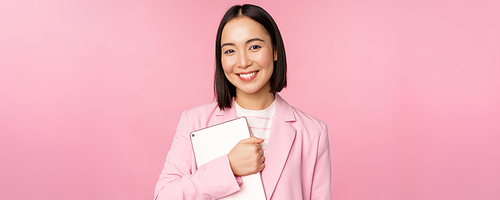 Image of young korean corporate woman, ceo manager holding digital tablet, smiling and looking professional, wearing suit, standing over pink background.