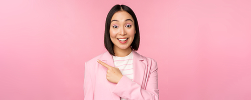 Portrait of asian business woman, saleswoman in suit pointing finger left, showing banner advertisement, smiling and looking professional, pink background.