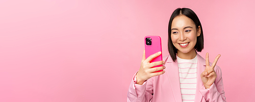 Stylish asian businesswoman, girl in suit taking selfie on smartphone, video chat with mobile phone app, posing against pink studio background.