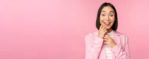 Happy young office lady, korean business woman wearing suit, looking surprised at camera, posing against studio background in pink.