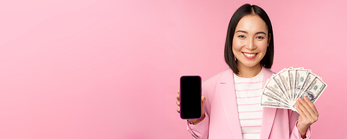 Image of korean successful corporate woman showing money, dollars and smartphone app screen, interface of mobile phone application, concept of investment and finance, pink background.