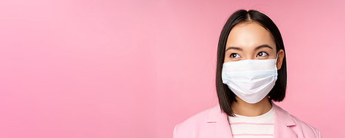 Close up portrait of japanese corporate woman in medical face mask from covid-19, looking left at logo, sale promo, standing over pink background.