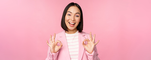 Okay, excellent. Businesswoman in corporate suit, showing ok, approval gesture, recommending smth, give positive feedback and smiling pleased, posing over pink background.