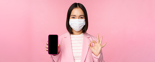 Portrait of asian businesswoman in medical face mask covid-19, showing smartphone screen and okay sign, pink background.