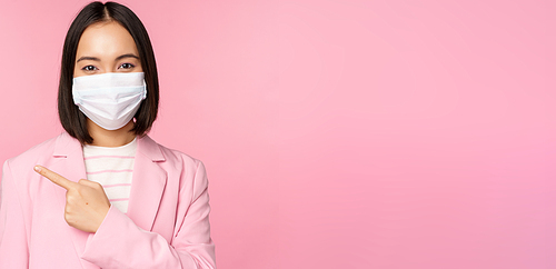 Portrait of asian businesswoman in medical face mask and suit, pointing finger left, showing advertisement, company banner, studio pink background.