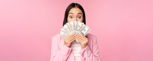 Happy asian lady in suit holding money, dollars with pleased face expression, standing over pink background. Copy space