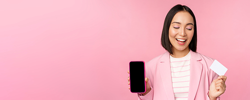 Smiling korean businesswoman in suit, showing mobile phone screen, credit card, showing online banking application interface, pink background.