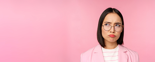 Disappointed asian businesswoman, office lady in glasses, looking upset at smth unfair, sulking, standing over pink background.